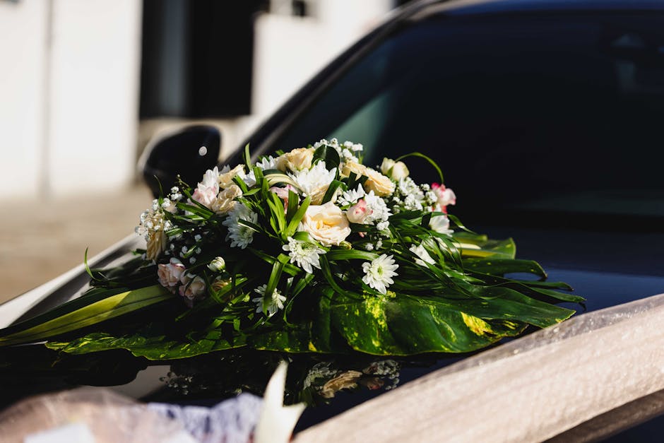 A delicate floral arrangement featuring white roses, daisies, and small pink buds, accented with lush green leaves, is beautifully placed on the hood of a black car. The bouquet is wrapped with large 