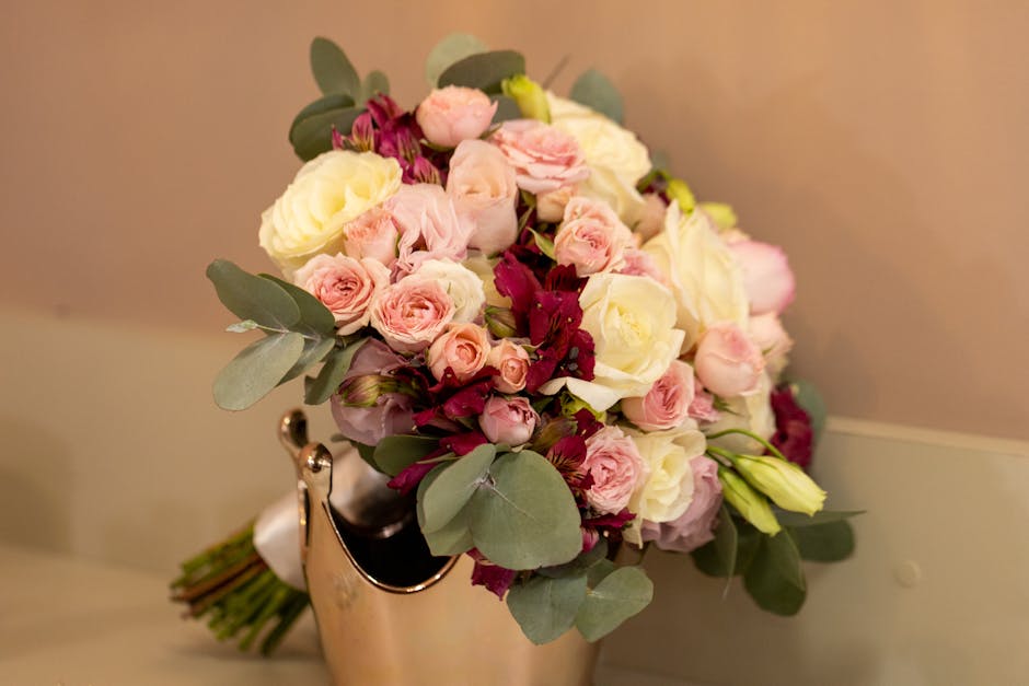 A bouquet arrangement featuring a variety of fresh flowers including soft pink and white roses, complemented by deep burgundy blossoms and lush green eucalyptus leaves. The bouquet is placed in a shin